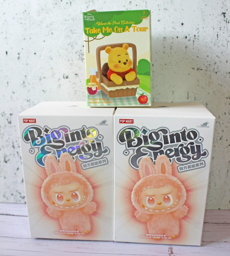 Three collectable boxes are displayed: one Winnie the Pooh-themed box labelled Take Me On A Tour sits on top of two larger boxes featuring a pink bunny character from the Big Into Persy series by Pop Mart.