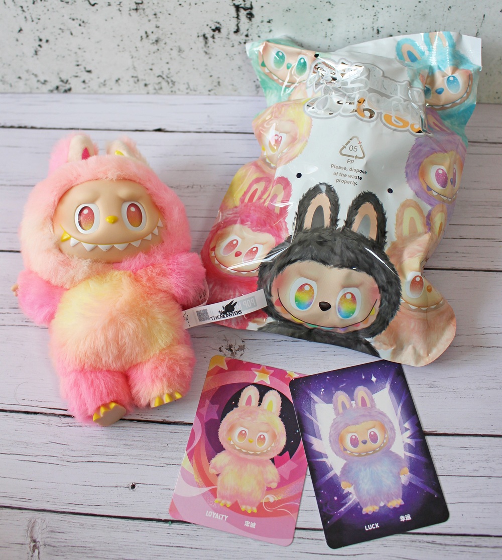 A soft toy with a pink, yellow, and white fluffy bunny suit sits next to colourful cards and its original packaging, featuring matching illustrations, on a white wooden surface.