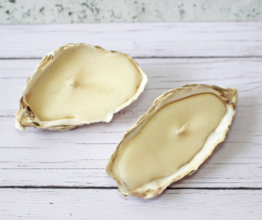Two oyster shells filled with creamy wax and wicks, resembling handmade candles, placed on a white wooden surface.