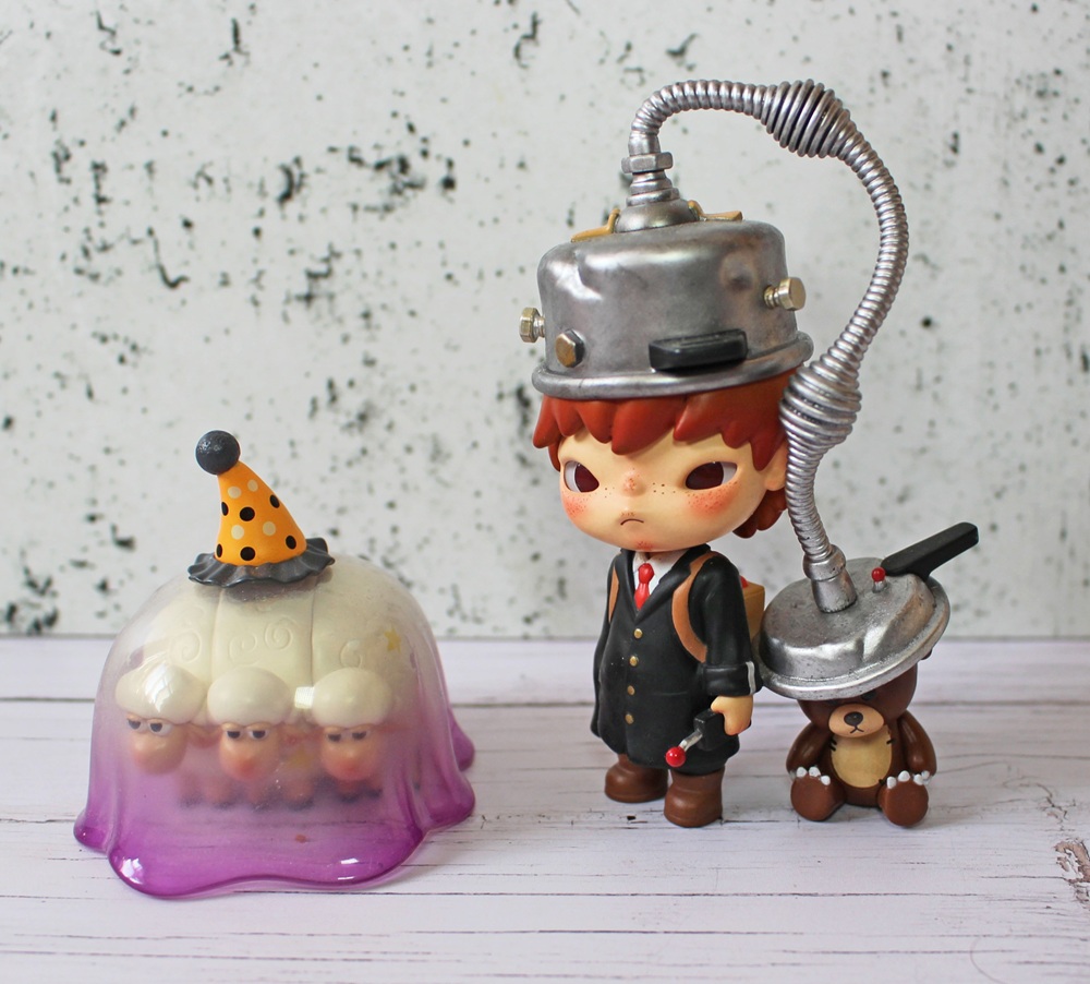 A toy figure with a metal helmet and suit stands next to a brown bear with a similar helmet. Nearby, four small figures in a purple jelly mould wear party hats, one orange with black dots. The background is white with black specks.