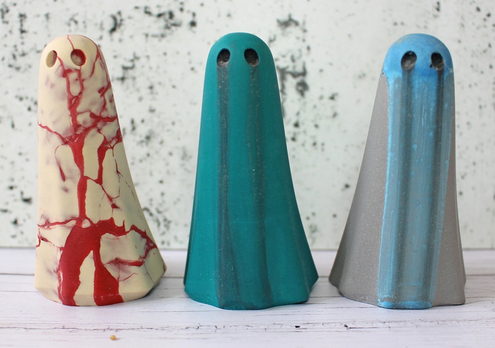 Three ghost-shaped ceramic figurines stand side by side. The left one is cream with red veining, the middle is teal, and the right is grey with blue accents. All have two small black eye holes.