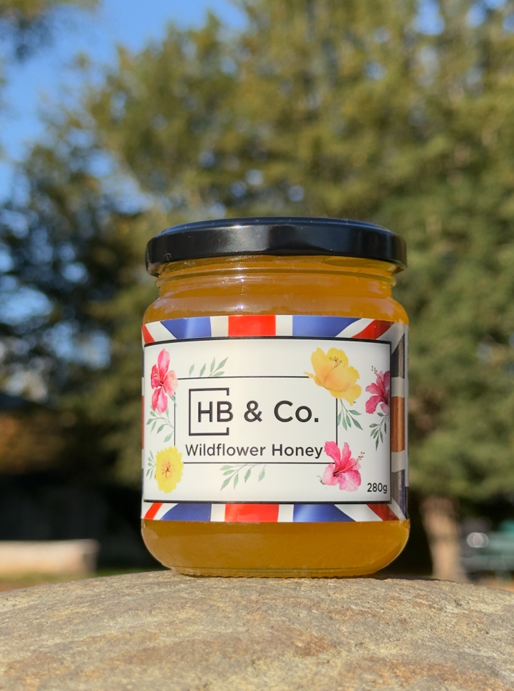 A jar of HB & Co. Wildflower Honey sits on a rock outdoors, with a label featuring colourful flowers and a Union Jack border. The honey is golden and the jar has a black lid. Trees are blurred in the background.