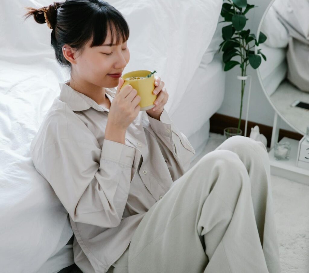 A woman in light-coloured pyjamas sits on the floor by a bed, eyes closed, holding a yellow mug with both hands, looking relaxed and content. A round mirror and a plant are in the background.