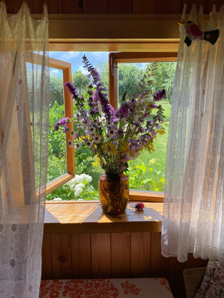 A vase of purple and white wildflowers sits on a windowsill with lace curtains, in front of an open window overlooking a sunny garden with green grass and trees. A small bird decoration is on the curtain rail.