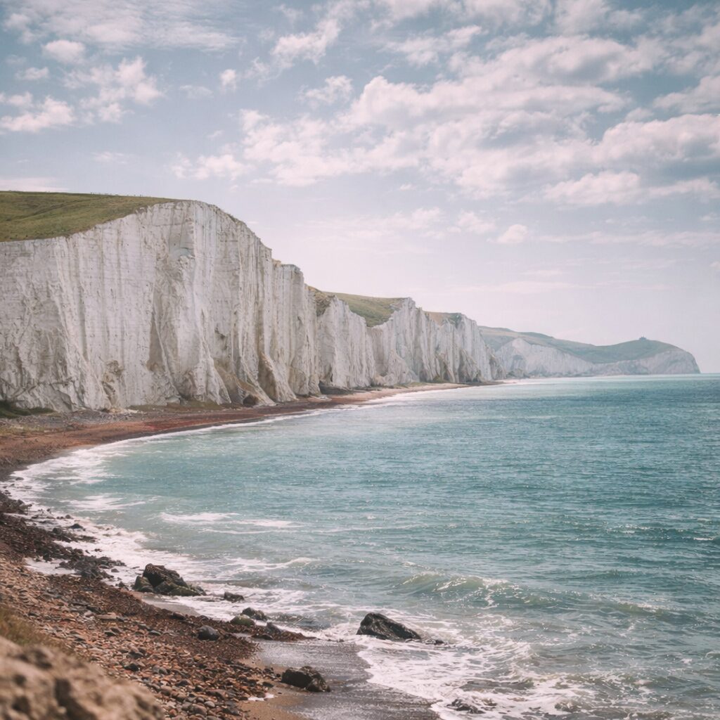 White chalk cliffs rise above a rocky coastline and calm blue sea under a partly cloudy sky, with green grass covering the cliff tops.