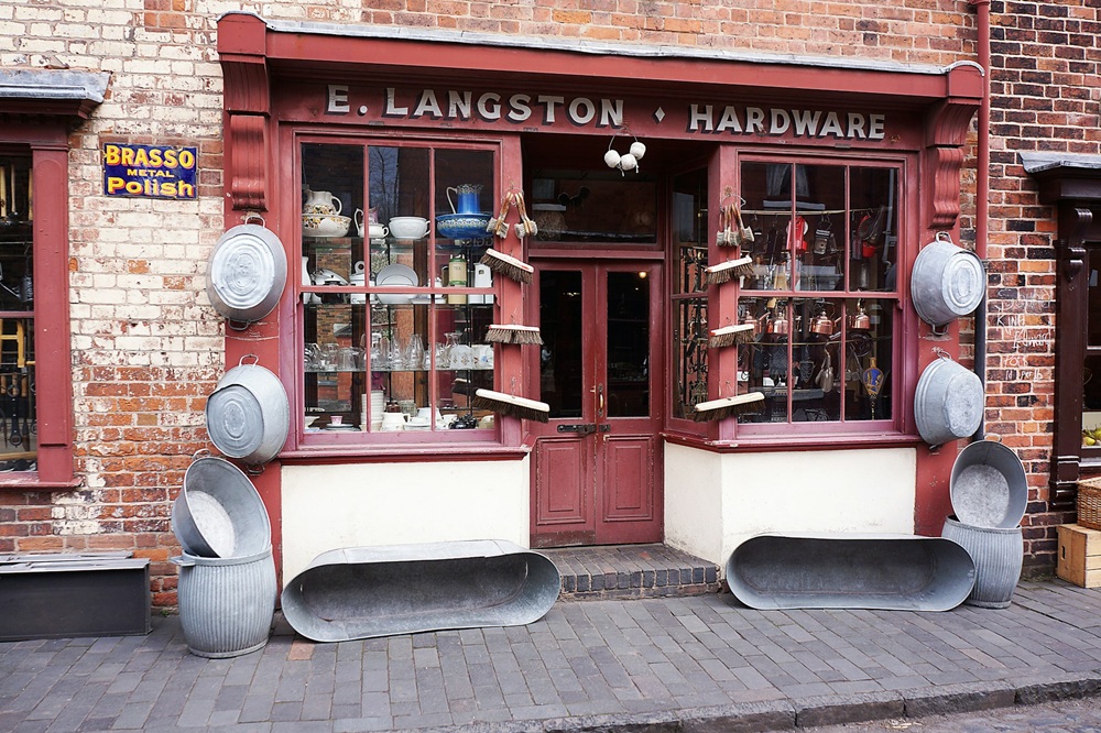 A vintage hardware shop with red trim, large display windows showing kitchenware, and metal tubs, buckets, and basins arranged outside. A sign reads E. Langston Hardware. A Brasso Metal Polish sign is on the wall.