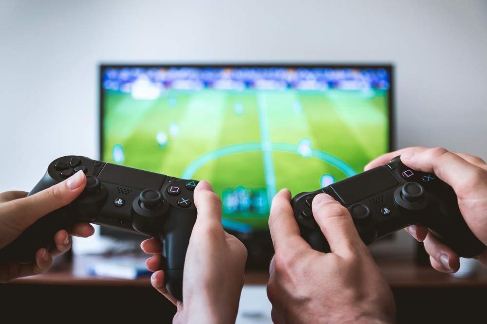Two people holding video game controllers play a football video game on a television, with the game visible in the background. The focus is on the controllers and players' hands.