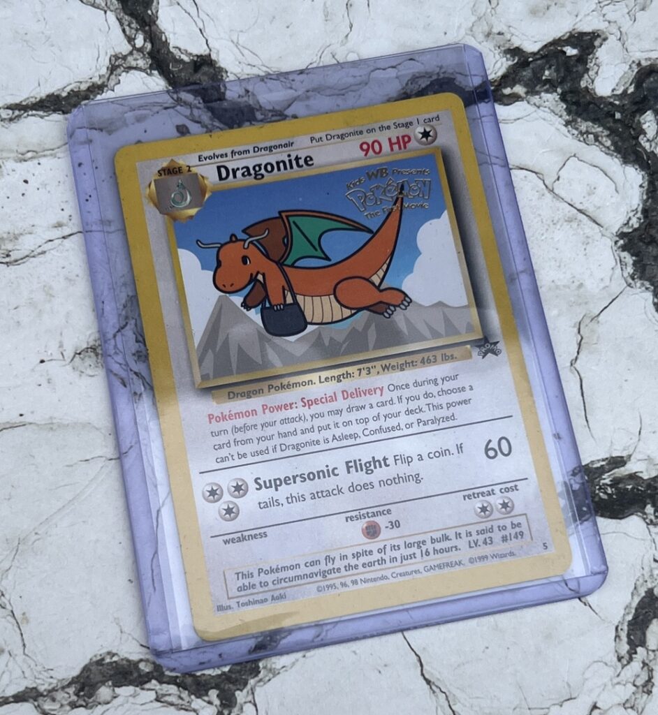 A Pokémon trading card featuring Dragonite in a protective case, placed on a cracked white surface. The card is titled Special Delivery Dragonite and shows a cartoon dragon with a postbag flying in the sky.