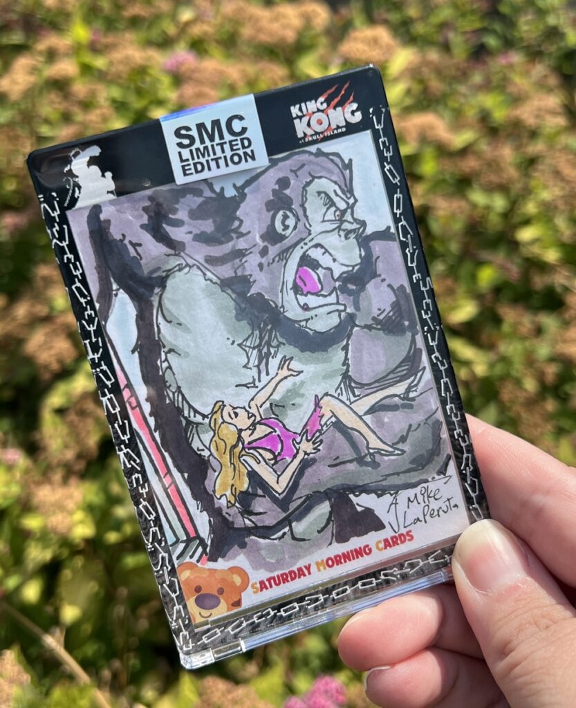 A hand holds a limited edition King Kong trading card featuring a cartoon drawing of King Kong holding a blonde woman. The card is signed Mike LaPeral and branded Saturday Morning Cards. Greenery is blurred in the background.