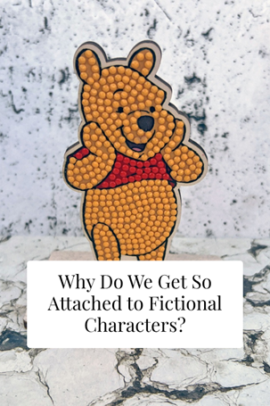 A cute bear figure in a red jumper stands with a happy expression. Text below reads: Why Do We Get So Attached to Fictional Characters? The background is a white, marbled surface.