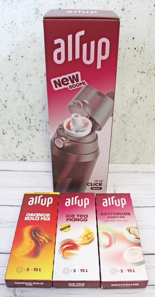 A red Air Up bottle box stands upright behind three Air Up flavour pod boxes: Orange Kola Mix, Ice Tea Mango, and Bestseller, displayed on a white wooden surface.