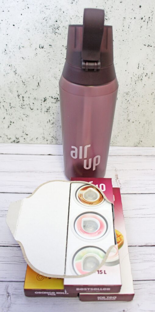 A purple Air Up water bottle stands behind an open box containing three circular flavour pods, displayed on a white wooden surface with a light speckled background.
