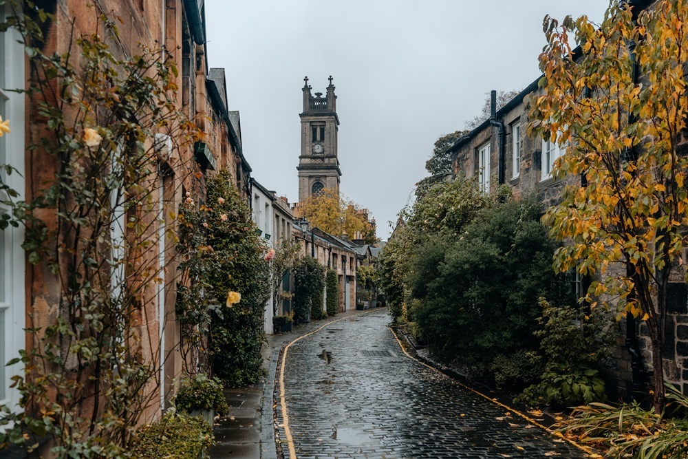 A narrow, cobbled street lined with stone houses and autumn trees leads to a tall church tower on a cloudy day.