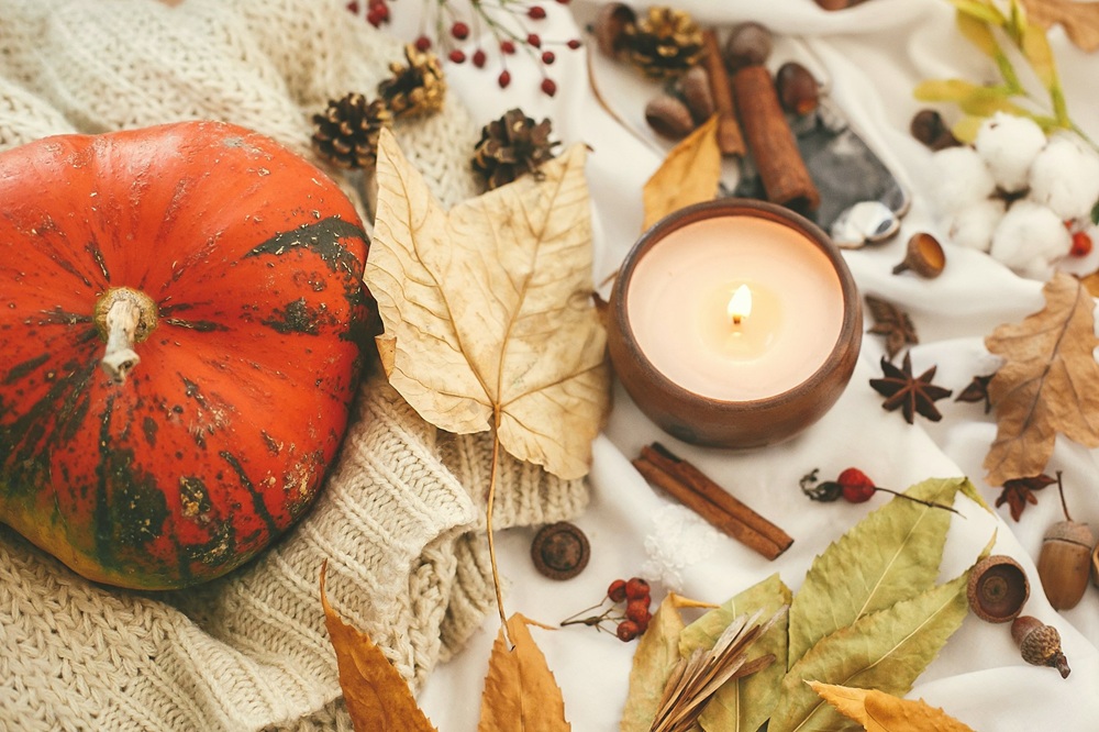 A lit candle, pumpkin, autumn leaves, pine cones, cinnamon sticks, and berries are arranged on a cosy knitted jumper and soft fabric, creating a warm autumn scene.