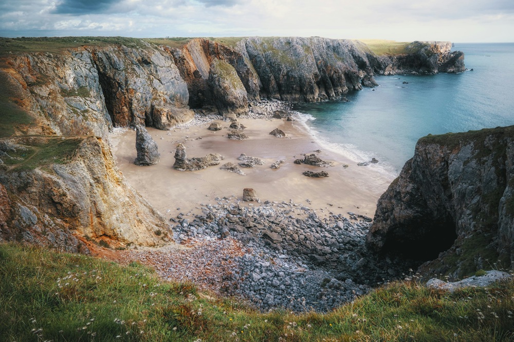 Rocky cliffs tower over a sandy beach scattered with large rock formations and pebbles, bordered by clear blue water under a partly cloudy sky. Grassy cliffs are in the foreground.