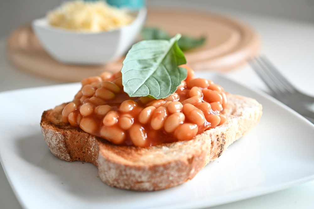 A slice of toasted bread topped with baked beans and a basil leaf on a white plate, with a blurred bowl of grated cheese in the background.